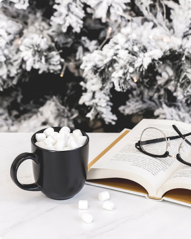 Holiday downtime for bloggers: warm cocoa, reading glasses, and an open book with snowy tree branches behind.