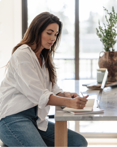 Woman journaling at a light wood desk with water nearby—symbolizing rewriting your story and starting fresh in business and life after 50. with the words "you can change the story" above.