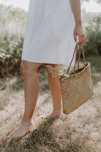 Woman walking barefoot through the grass carrying a weaved bag with wild flowers. Savage Daughters