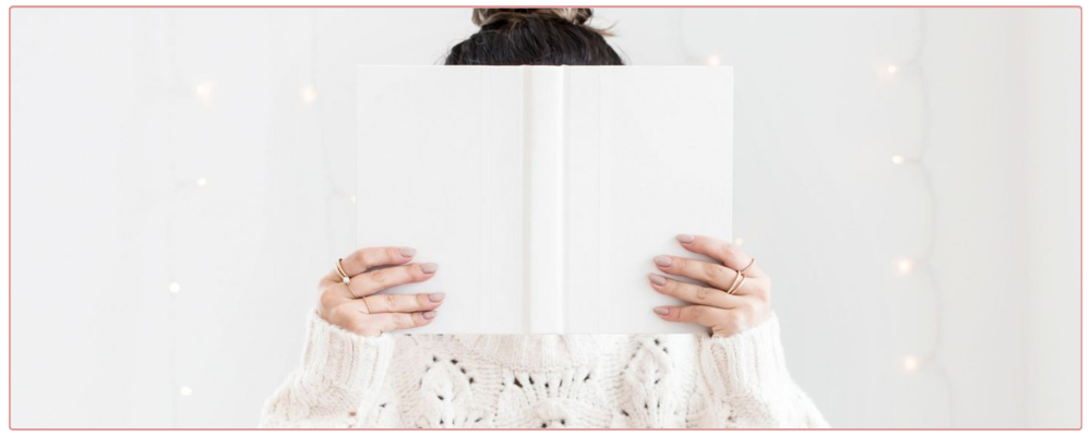 Woman hiding behind a book, wearing a white sweater with twinkle lights behind her