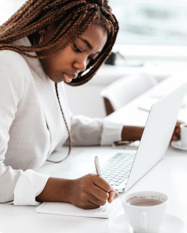 woman coach writing blog post notes beside laptop and coffee while doing a 10 minute blog review of her coaching blog.