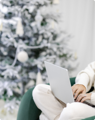 Cozy holiday blogging scene with a woman typing a blog post on her laptop beside a softly lit Christmas tree, capturing calm holiday blog ideas.