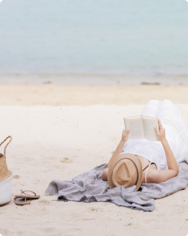 Woman in sunhat reading on the beach with sandals and a tote nearby—evoking calm, quiet marketing, and sustainable summer business energy.