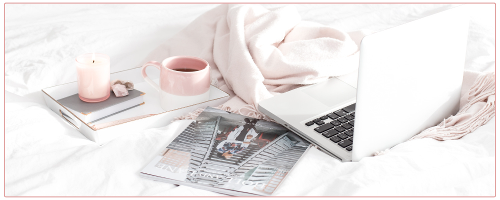 laptop, tea in a pink mug, and magazine on a white bed with a pink throw blanket. 