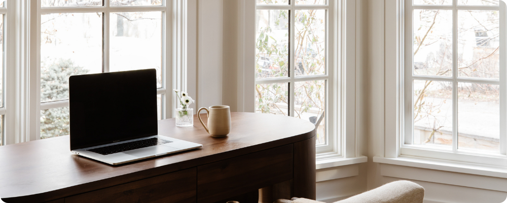 Wide desk with laptop and mug by bright windows, blogger setup for weekly blogging routine and blog ideas.