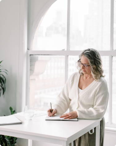 Woman writing in a notebook by a bright window, reflecting on her Stop-Doing List to simplify business and reduce overwhelm.