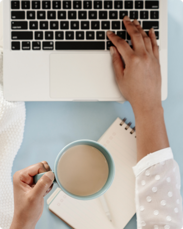 Person typing on laptop while holding coffee mug, planning blog posts for coaches with calm, focused energy.