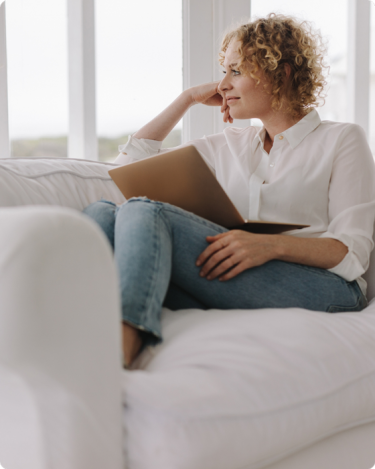 Woman gazing out the window, with laptop on her lap, sitting on a couch—visual for blog post on how to say no without guilt and set boundaries as an introverted coach or solopreneur.